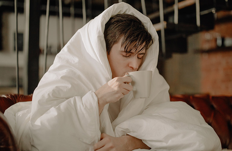 man with duvet on head drinking from a cup